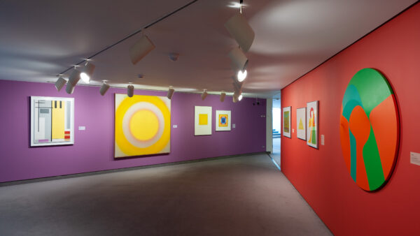 Gallery image. On the left is large abstract painting in yellow prominently displayed on a bright purple wall. On the right is a red wall with several paintings