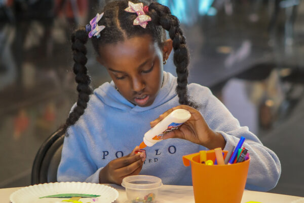 Young African American girl, her hair in two braids and wearing a purple, long-sleeved shirt. She's sitting at a table, holding a bottle of glue in her hand and applying glue to an object in her other hand. Various art supplies are on the table front of her.