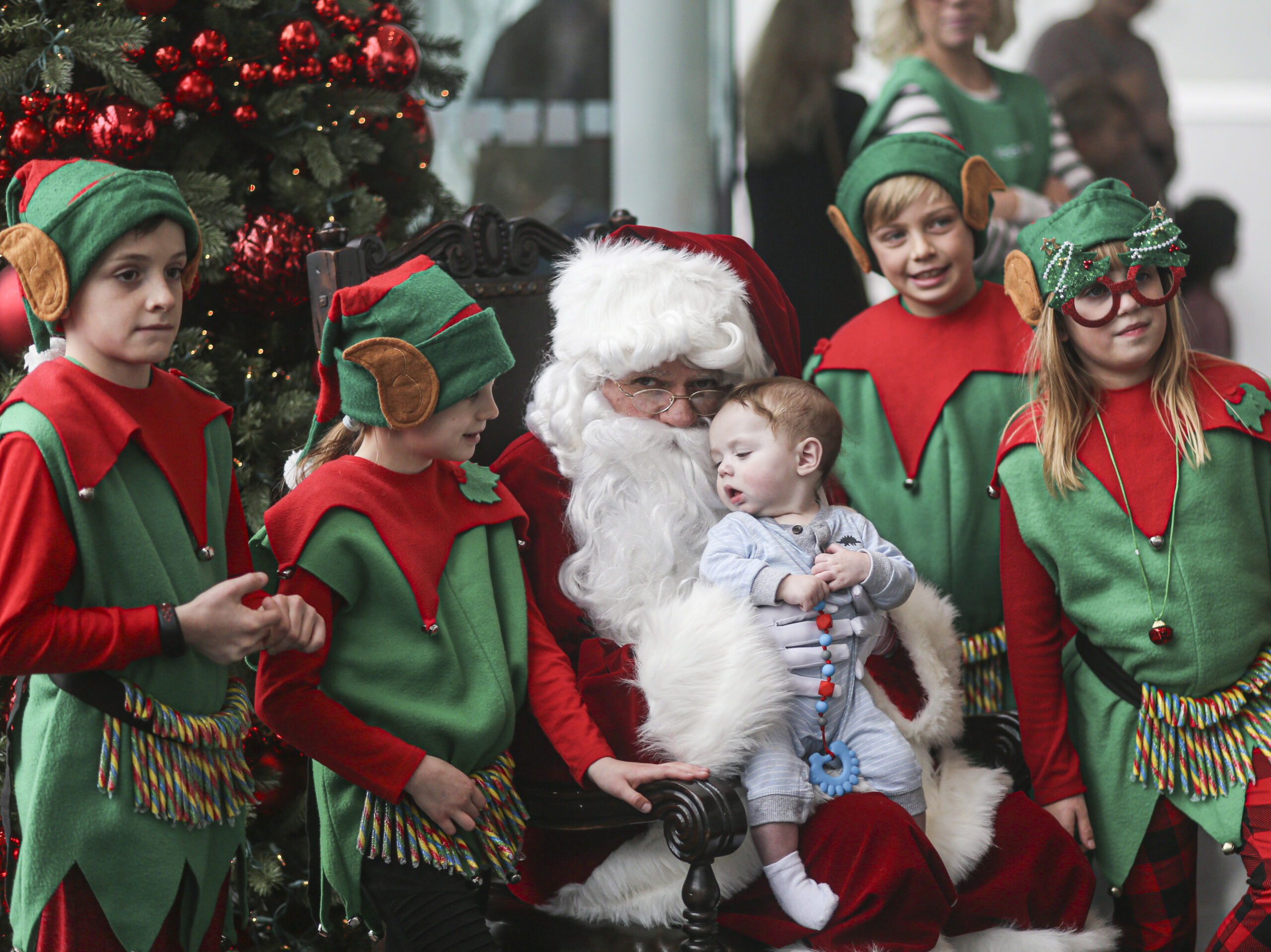 Santa Claus, seated in front of a Christmas tree, holding a baby, with children in elf costumes of red and green standing around.