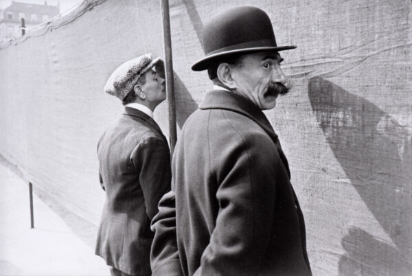Two men standing facing a wall. Man on the left wears a newsboy-style cap and is dressed in a dark jacket. Man on the right wears a black bowler hat, has a dark mustache and is dressed in the dark jacket.