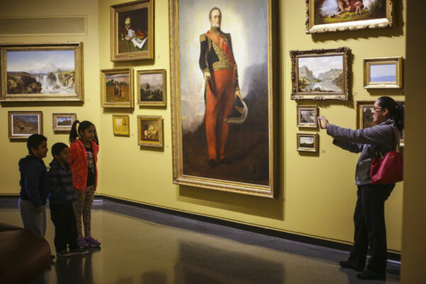 Family in the gallery. Three children stand in a group looking at a portrait on the wall, with an adult standing opposite, holding out a cell phone to take a photograph