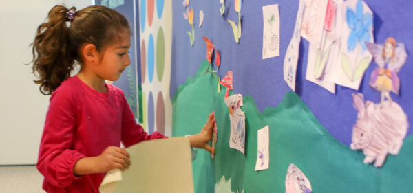 Young girl with dark hair pulled into a ponytail, dressed in a long-sleeved red shirt, stands in front of a wall with a background of the curvy green border at the bottom and a blue background on top with pieces of children's artwork displayed. The girl holds a piece of paper in one hand, with the other outstreched toward the wall