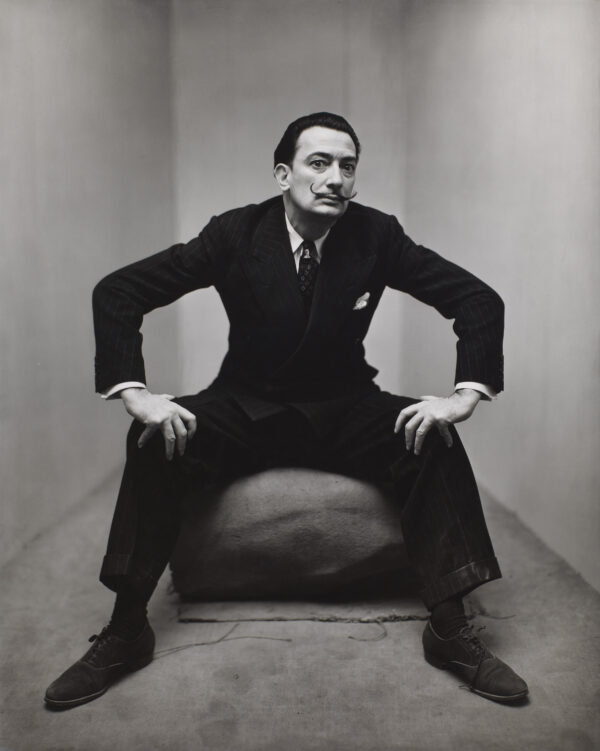 Photograph of Salvador Dali, seated with knees apart, hands on knees, dressed in a dark suit.