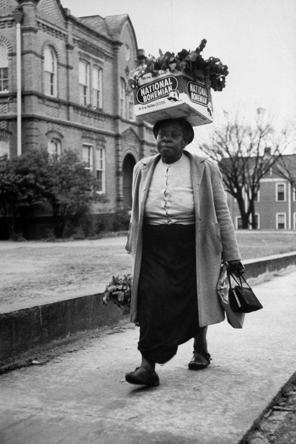 Photograph of an African American woman walking with a box of groceries on her head. She's dressed in a gray coat, white button-up blouse and dark skirt. She carries a bag of groceries in her right hand and a another bag and black handbag in her left hand. A multistory brick building is in the background.