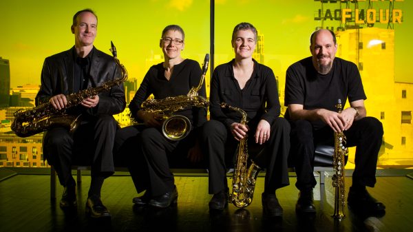 Four peop;e seated on a low bench in front of a yellow background. All four hold saxophones
