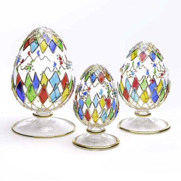 Three glass eggs, mostly clear glass with diamond shapes of shades of blue, red, yellow, and green of varying sizes. Sitting on a clear base with a round foot and gold trim