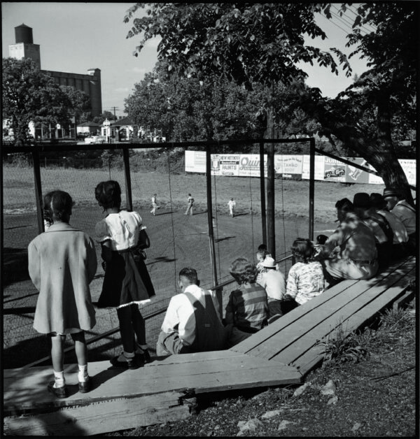Photograph of two African American children standing at a fence overlooking a baseball diamond. At right are people seated on bleachers.