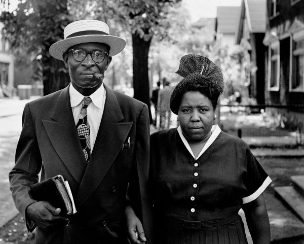 African American man and woman walking outside. Man, on the left, dis dressed in a dark suit, white shirt, and dark tie, wearing a white boater hat. He is smoking a cigar and carries a book in his left hand. Woman on the right is dressed ina dark top, with white around the v-neck and short sleeves. She wears a net hat.