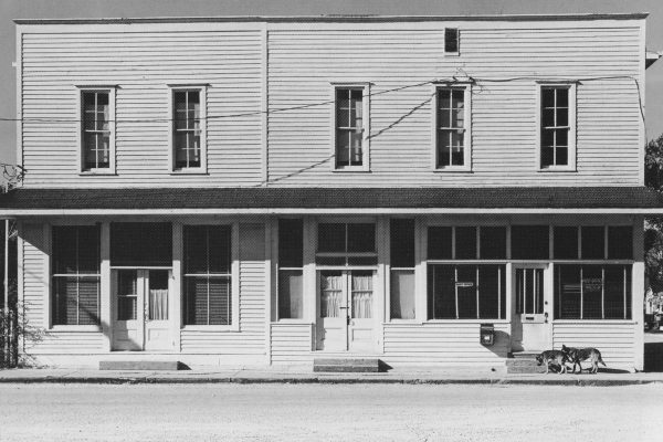 Photograph of white two-story wood building with a porch along the front. Five tall narrow windows are along the top floor of the building. The lower level had wider windows and doors. Two dogs are in front of the building on the right side of the image