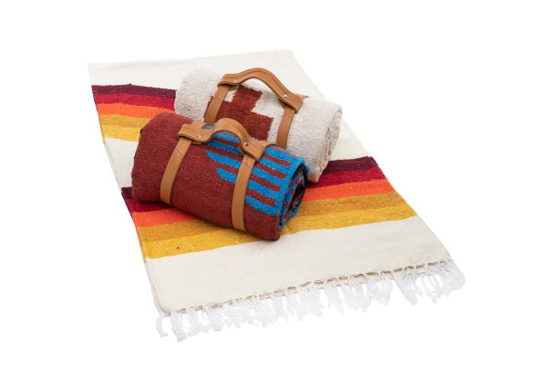 Red blanket with a blue geometric patter, rolled and secured with a lightbrown leather-like belt and a white blanket with a dark red cross, sitting on an unfolded blanket of white with stripes of yellow, gold, orange, and brown