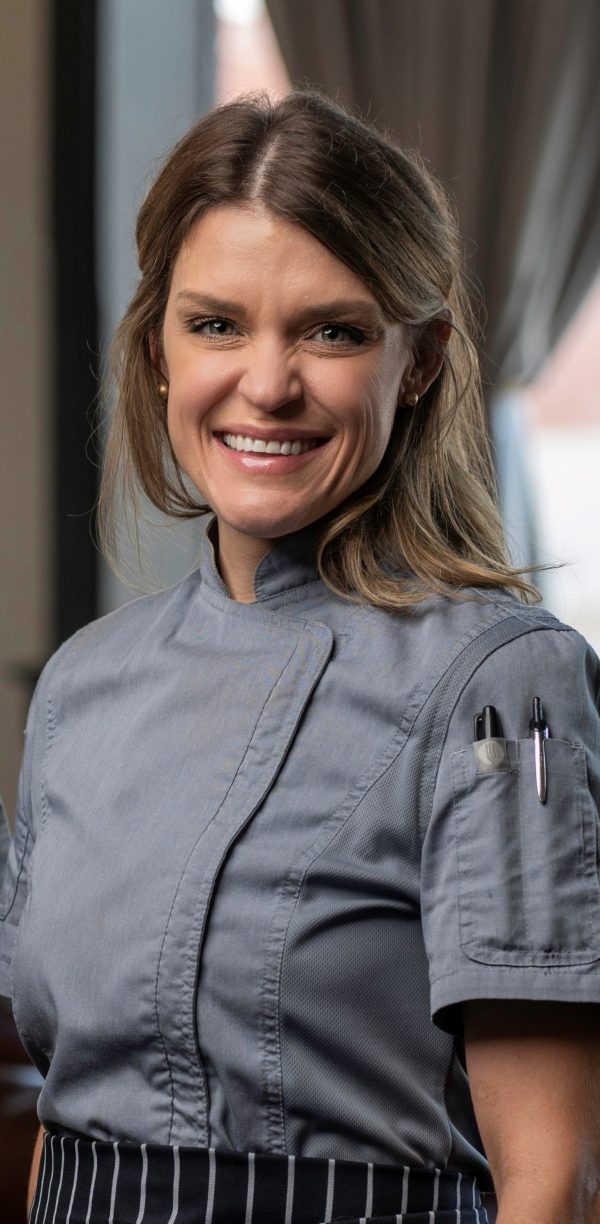 Headshot of a white woman with light brown hair, smiling for the camera and wearing a grey chef's jacket