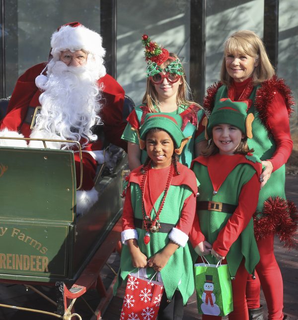Santa Claus sitting in a green sleigh. Three girls dressed in red and green elf costumes, and a woman, also dressed in red and green are standing beside him